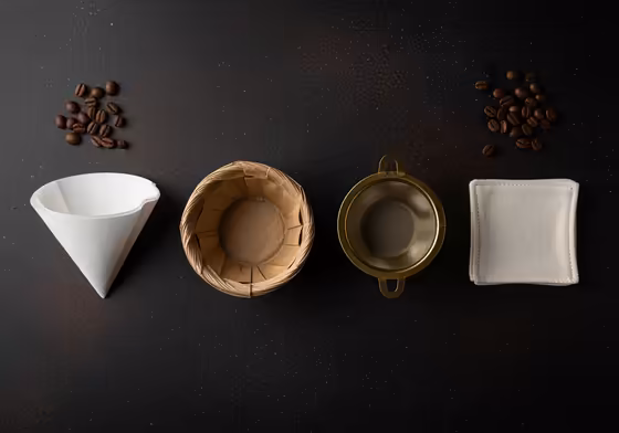 A collection of different coffee filters including paper cone, paper basket, metal mesh, and cloth, arranged on a rustic wooden surface.