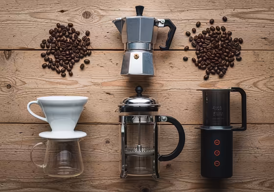 A collection of different manual coffee brewing devices like a French press, pour-over dripper, AeroPress, and Moka pot arranged on a wooden table.