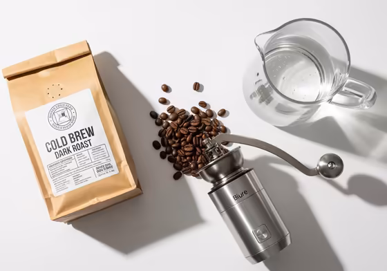 A flat lay of premium cold brew ingredients: a bag of fresh, dark roast coffee beans, a high-quality burr grinder, and a glass pitcher of filtered water.