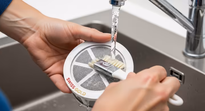 A person rinsing a Swiss Gold filter under a kitchen tap with a soft brush to clean it.