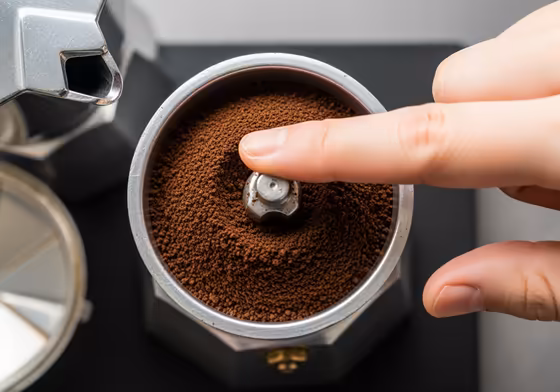 A close-up view of a moka pot's filter basket being filled with medium-fine coffee grounds, leveled off with a finger.